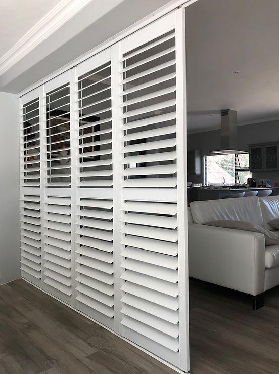 White bi-fold shutters stacked as a room divider between a lounge and kitchen